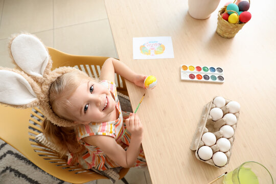 Cute little girl in bunny ears painting Easter eggs at home
