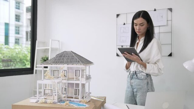 Beautiful caucasian architect focus on using tablet to check house model on meeting table with blueprint, house model and architectural document scatter around at meeting room. Immaculate.