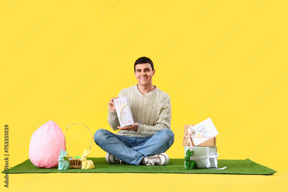 Wall mural Happy young man in bunny ears with gift boxes and Easter eggs on yellow background