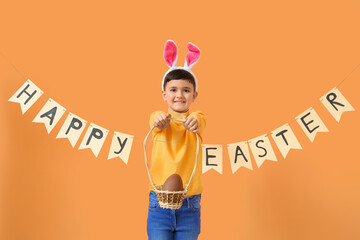 Cute little boy in bunny ears headband holding basket of chocolate egg and paper garland with text...