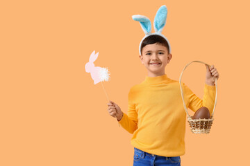 Cute little boy in bunny ears headband holding basket with chocolate egg and decor for Easter on orange background