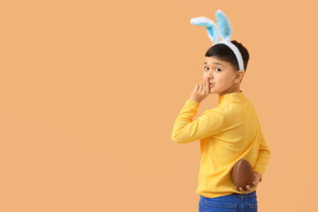 Cute little boy in bunny ears headband with chocolate egg for Easter showing silence gesture on...