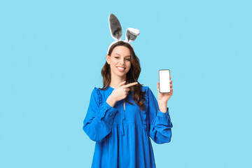 Happy young woman in Easter bunny ears headband pointing at mobile phone on blue background