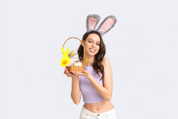 Happy young woman in bunny ears headband holding basket with flowers and Easter eggs on white background