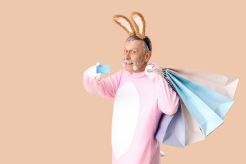 Senior man in Easter bunny costume with shopping bags and credit card on beige background