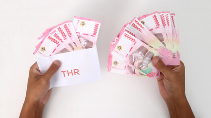 Hand holding a THR envelope filled with Indonesian Rupiah banknotes. THR or Tunjangan Hari Raya is...