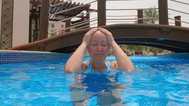 Lockdown shot of woman in clear water swimming pool looking camera. Female enjoys in water at resort. She strokes her face and hair. She is spending leisure time during vacation. Bridge background.