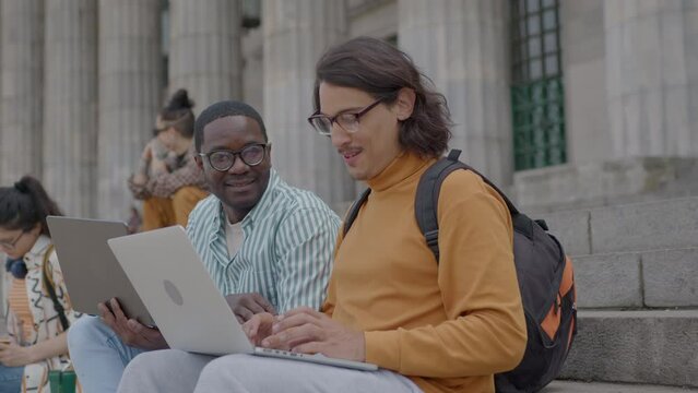 Young Latin male student discussing homework with African-American groupmate and using laptop when sitting together on stairs outside the university