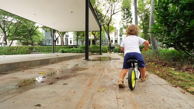 Bold toddler on runbike speeds up to ride through puddle, but slips on wet surface. Mishap unfolds in slow motion as young child loses control, experiencing tumble with minor scrape.