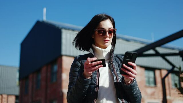 Frustrated woman in stylish glasses using mobile gadget with credit card in hand while standing on street. Angry woman opening online banking application and noticing absence of salary enrolment.