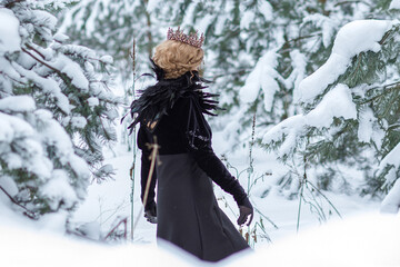 Fototapeta premium Fashion concept. A beautiful young lady with blond hair and a long black dress, with a crown on her head, stands in a winter park. Beautiful dark princess using a black magic spell.