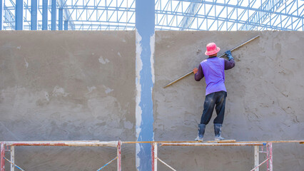 Asian builder worker on scaffolding is using long trowel to plastering cement on concrete wall of industrial building in construction site, rear view with copy space