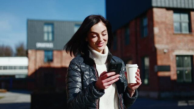Beautiful woman in jacket typing on mobile phone touch screen while having stroll with takeaway cappuccino. Remote freelancer going out for coffee break while working day and checking mailbox.