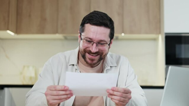Smiling young man holding paper bill letter while working at table with portable laptop indoors. Skilled caucasian freelancer feeling shock while reading recieved information at home interior.