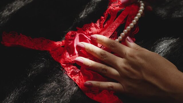 A woman's hand touches red lingerie with white pearls. The image has a sensual and intimate mood.