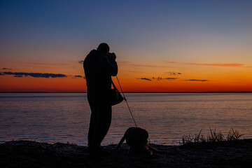 Silhouette of a guy with photo camera and dog on a river shore at sunset