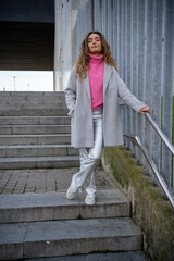 young blonde woman looking forward wearing a gray coat and a pink sweater with silver pants