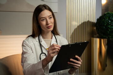 A focused young woman in a white lab coat, with a stethoscope around her neck, is writing notes on a clipboard. She appears to be a medical professional, probably a doctor or a nurse, standing in an