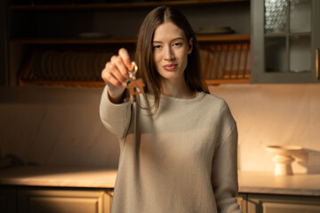 A cheerful young woman stands in a dimly lit modern kitchen, holding up a set of keys towards the camera, symbolizing a new home or ownership. Her casual attire and subtle lighting suggest a
