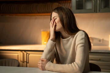 A young woman is depicted sitting at a kitchen table, her body slightly hunched in a pose that...