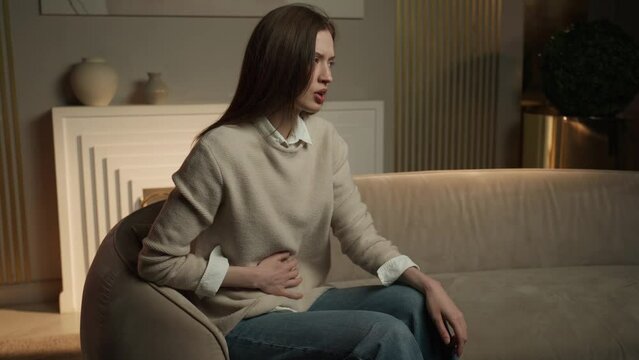 A woman is seated on a sofa clutching her abdomen with a pained expression, suggesting she is experiencing stomach discomfort. The setting appears to be a cozy living room, and the situation indicates