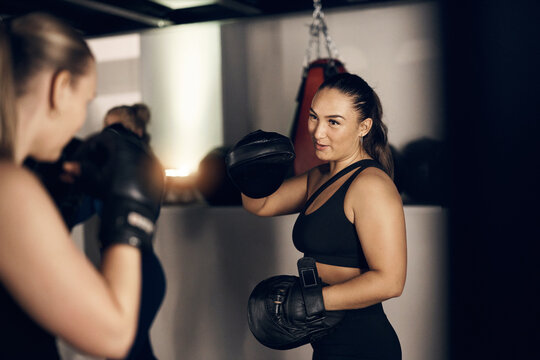 Smiling young woman wearing a tank top and hand pads during punching practice with a female friend during a boxing session together at the gym