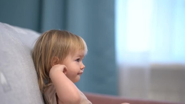 Cute kid toddler looks at the TV and smiles, focused gaze behind the camera. 4k video.