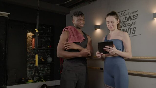 Professional attractive female fitness instructor and handsome muscular build African man friendly talking and using digital tablet for schedule exercise training, while standing at gym after workout.