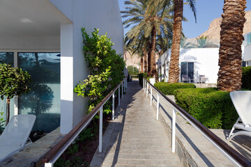 Walkway surrounded by lush greenery and white building with its reflective glass windows. Palm trees tower above casting gentle shadows on the path under the blue sky. A stone walkway with railings