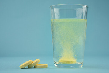 Photograph of orange colour water soluble vitamin C dissolving in glass of water with bubbles and pile of pills or tablets on blue colour background