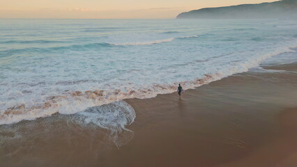 Lonely surfer entering ocean drone shot. Wonderful pastel sky with marine waves