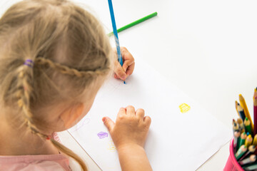 A child blonde girl draws with colored pencils sitting at the table. Top view, flat lay.