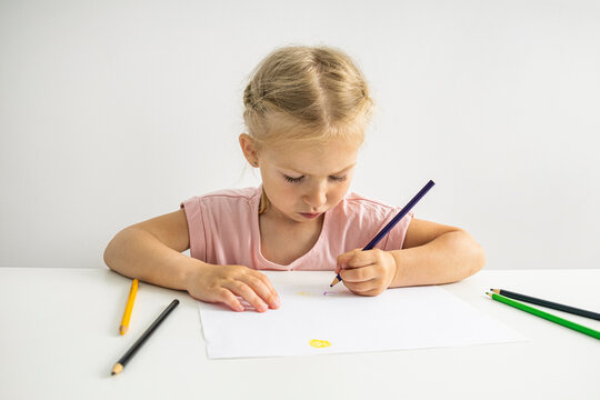 The girl draws with colored pencils sitting at a white table on a white background