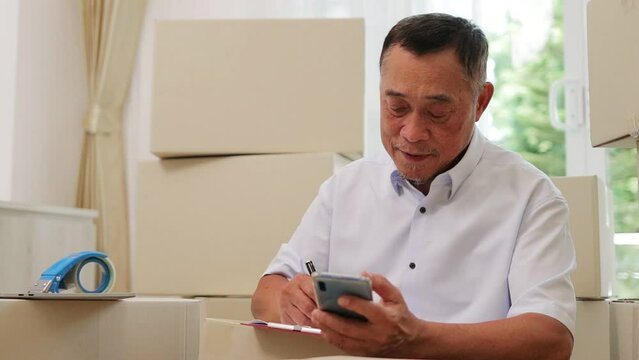 An elderly Asian man running an online business is checking information on his smartphone in preparation for sending items to customers. SME business. Entrepreneur. Working in retirement