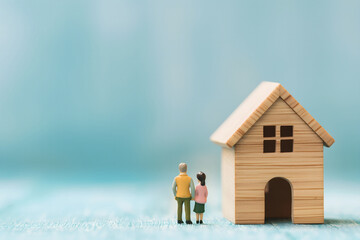 Two miniature people are standing in front of wooden house, light blue background