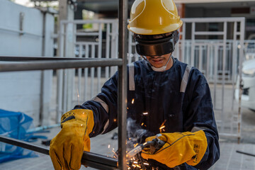 Asian worker wearing goggles, gloves, hat and safety suit uses a welding machine. Sparks from...