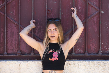 Fototapeta na wymiar Young, beautiful blonde woman in black skirt and black top is raising her arms and holding a fence of a house. The young woman looks at the camera with a serious face.