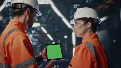 Engineer teaching woman intern on factory showing green screen tablet close up.