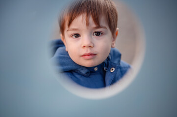 A charming toddler peeks through a playground tunnel, highlighting the curiosity and delight of a child at play against a muted background
