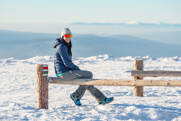 Young woman sits on a wooden stump on top of a mountain and rests after hiking on a winter mountain trail, mountain landscape in winter.