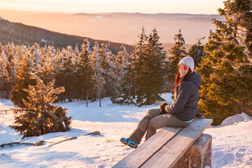 A young woman sits on a bench in the mountains and rests after hiking on a winter mountain trail.
