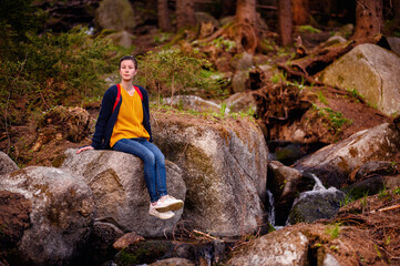 Amidst a rustic forest setting, a young woman finds a moment of reflection while seated on a moss-covered boulder