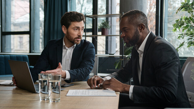 Diverse multiracial African American and Caucasian men colleagues coworkers businessmen financial agents discuss business project startup with laptop in office work with papers documents paperwork