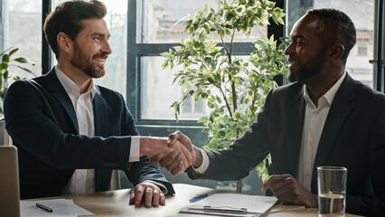 Two diverse multiracial businessmen signing document agreement shaking hands success business deal. Multiethnic men partners in office meeting handshake satisfied agree sign loan investment contract