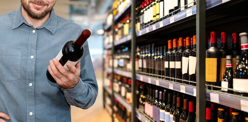 man consumer chooses a wine in supermarket. Man holding wine bottles in hand and smiling in liquor store.