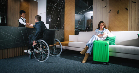 Man in wheelchair checking in with help of lovely attractive professional female receptionist ready...