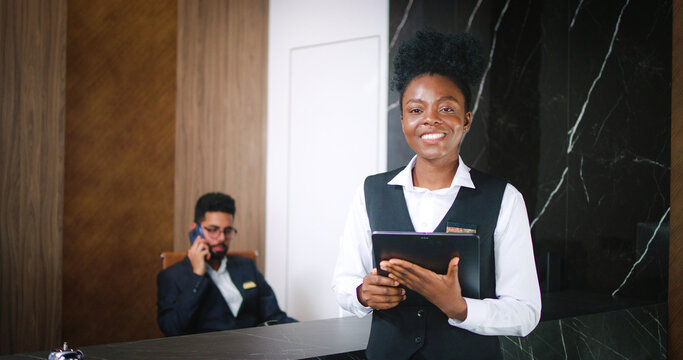 Portrait of beautiful young professional receptionist using tablet online looking at camera and smiling. Busy luxury hotel manager talking on phone taking calls on background.