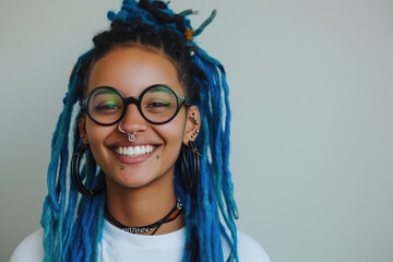 A joyful woman with blue dreadlocks and a septum piercing wearing round glasses smiles gently against a neutral background.