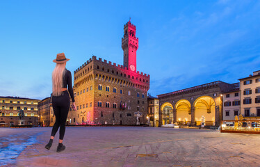 Fototapeta premium A beautiful blonde woman with a stylish hat wearing black tight trousers walking on the street - Palazzo Vecchio or Palazzo della Signoria at twilight blue hour - Florence, Italy. 