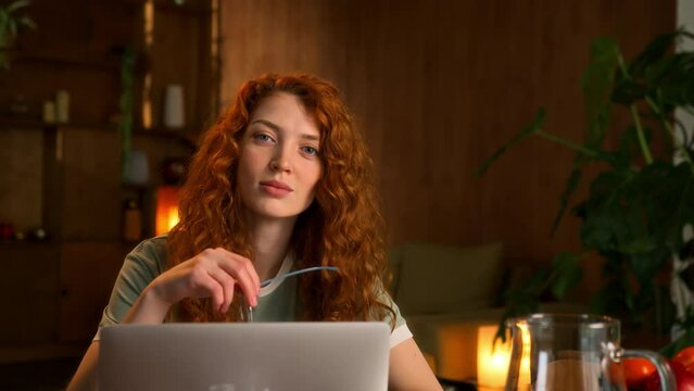 portrait of young red haired confident and contented woman sitting at table working at laptop at home. takes off eyeglasses, looks in camera smiling